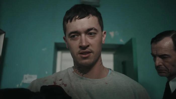 Wasteman: Tom Blyth stands with blood on his prison clothes while a guard stands behind him