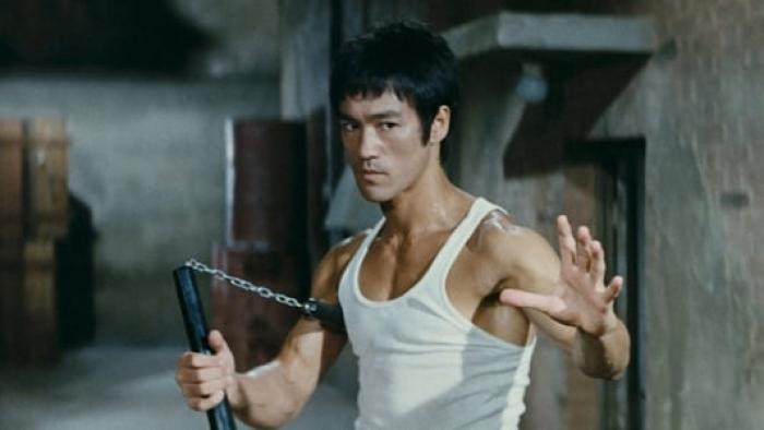 The Way of the Dragon movie: Bruce Lee in warehouse wearing white vest holding nunchucks.