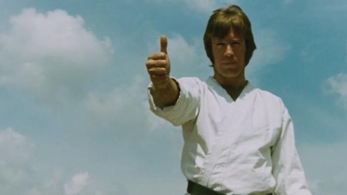 The Way of the Dragon movie: Chuck Norris stands in white kung fu uniform giving thumbs-up with a blue sky background.