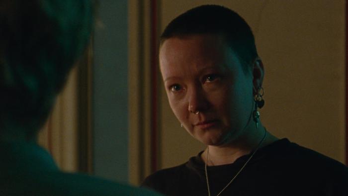 Blue Jean: A White woman with a short shaved haircut and a ring through her nose talks to someone not shown 