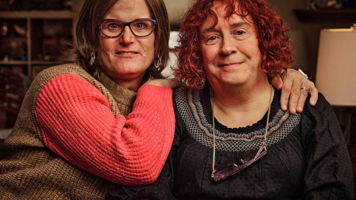 Still from We Live Here: The Midwest: Seated and facing the camera are Jenn Richmond (left) and Debb Richmond who are a White LGBTA+ couple from Minnesota, wearing casual attire