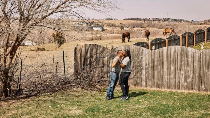 Still from We Live Here: The Midwest: Embracing each other while standing on farmland in Nebraska are Mario Foreman-Powell (left) and Monte Foreman-Powell, a Black and married LGBTA+ couple, wearing casual attire 