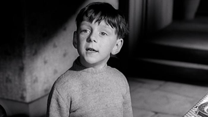 Whistle Down the Wind: A young White boy stands by table in front of stairs