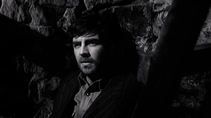 Whistle Down the Wind: A White middle-aged bearded man sits on straw against a wall in a barn while wearing a suit