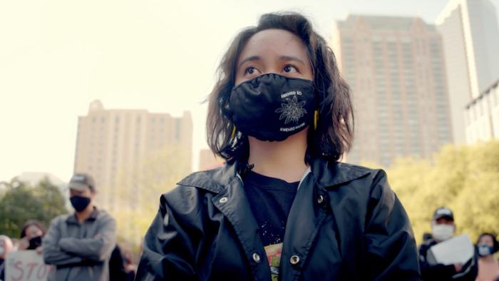Still from Who We Become: Filipino community organizer Jenah Maravilla looks up at the stage during a gathering of a diverse group of people at a stop Asian hate crime vigil and rally in Houston, Texas, wearing a black windbreaker jacket, a dark-colored graphic top, and a black face mask
