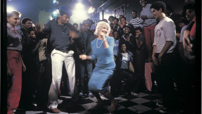 Still from Wild Style: White newspaper reporter Virginia (Patti Astor) with neck-length straight bleached-blond hair dancing at a club and surrounded 