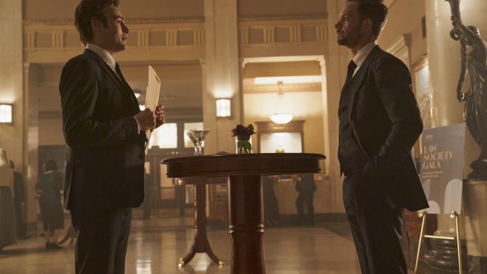 Young Werther movie: Douglas Booth and Patrick J. Adams stand across from each other in a fancy event space hallway, wearing suits