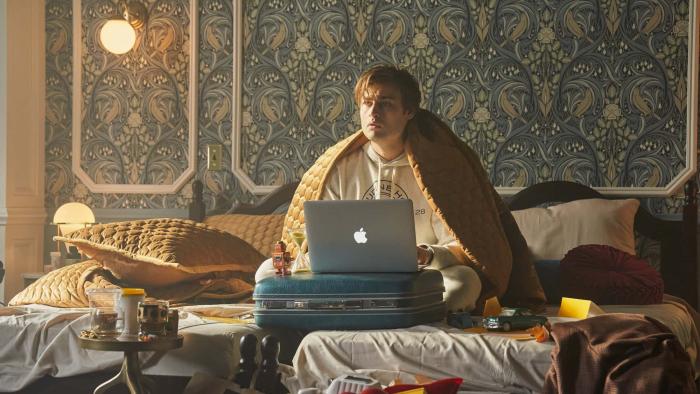 Young Werther movie: Douglas Booth wears a blanket over his shoulders, sitting on a hotel bed with a laptop