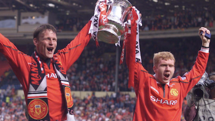 99 TV show screenshot 2: Two Manchester United players in red jackets hold up trophy. 