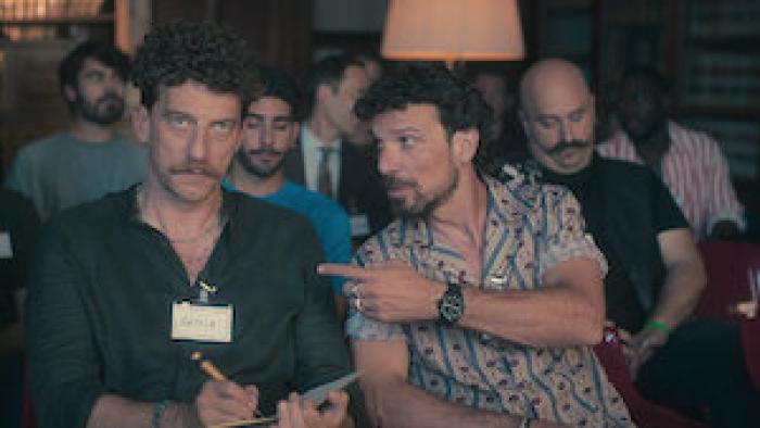 Real Men TV show screenshot: A White man points at another White man who's taking notes in a seminar with more men in the background