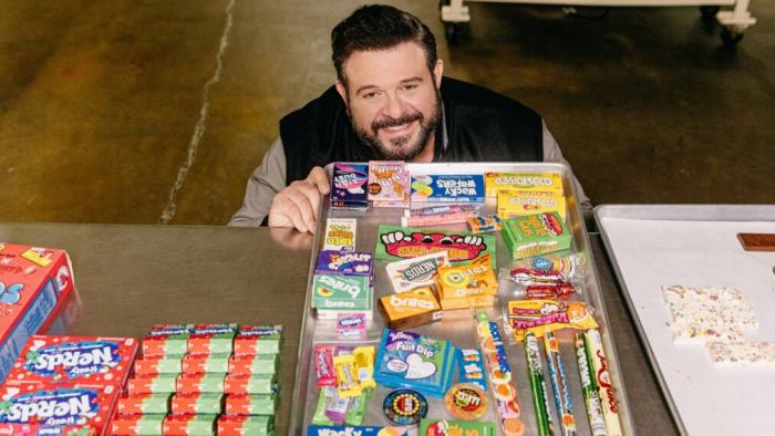 Adam Eats the 80s: A white man squats behind a table full of 80s candy like nerds