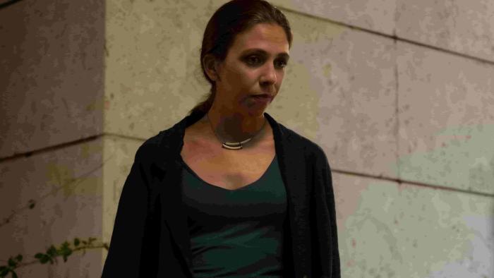 Adoration TV show screenshot 3: Barbara Chichiarelli stands in front of a stone wall. She wears a blue shirt and black blazer.