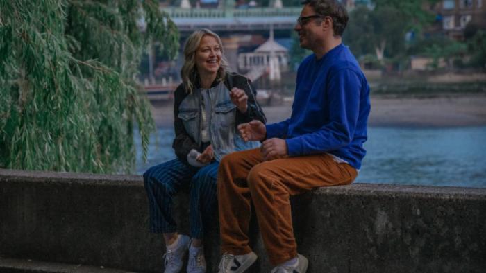 Alice and Steve TV show screenshot 1: Alice and Steve sit on a cement wall next to a river; they are smiling at each other and gesturing broadly
