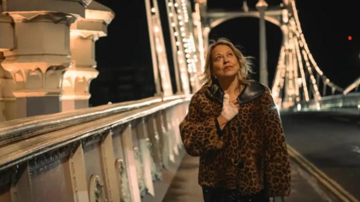 Alice and Steve TV show screenshot 3: Alice walks underneath a lighted bridge wearing a leopard print coat; she smiles up at the sky
