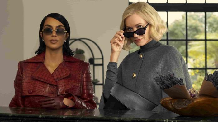 Kim Kardashian and Naimi Watts at a counter. Kardashian wears a red leather coat. Watts wears a dark grey top with buttons.