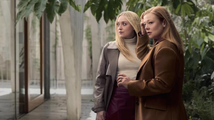 A blonde and redheaded woman stand together outdoors wearing business attire looking in different directions with concern.