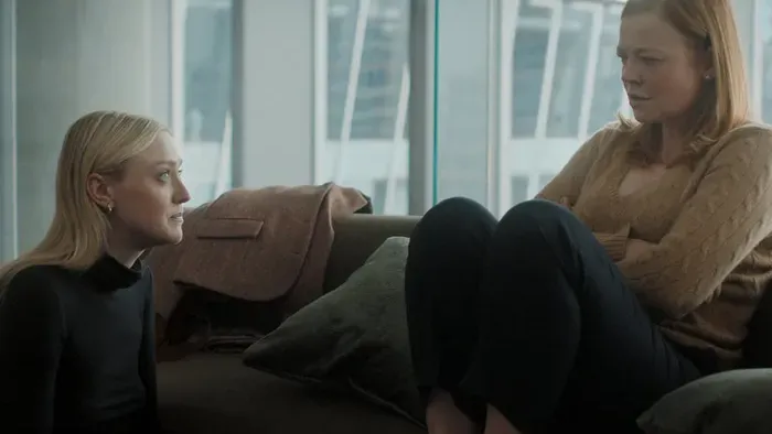 A blonde woman sits on the floor next to a couch and looks up at a professional looking redheaded woman who has her shoes off and feet on the couch.