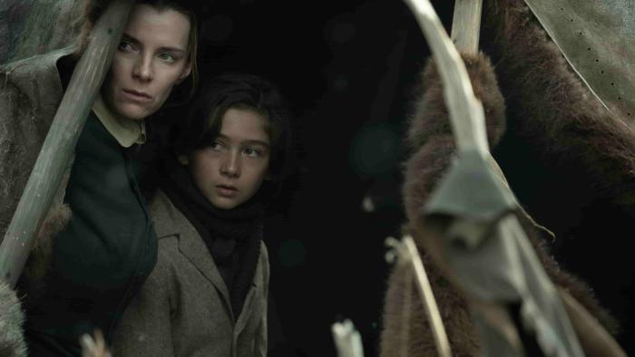 American Primeval TV show screenshot 1: Betty Gilpin and Preston Mota hide in a tent. Gilpin wears a black dress, and Mota wears a grey coat.