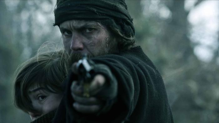 American Primeval TV show screenshot 2: Taylor Kitsch holds Preston Mota to his chest as he aims a gun. He wears a black coat, fingerless gloves.