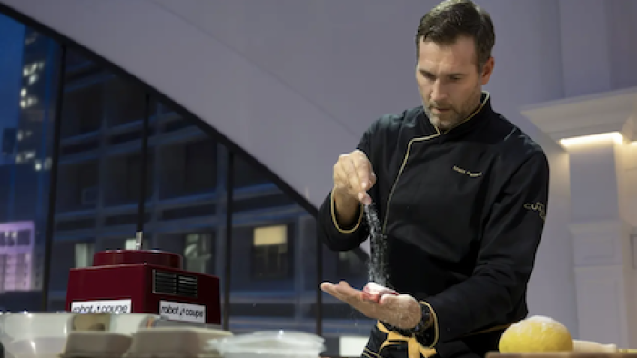 America's Culinary Cup TV show screenshot 2: A contestant wearing blue chefs coat seasons something in his hand. 