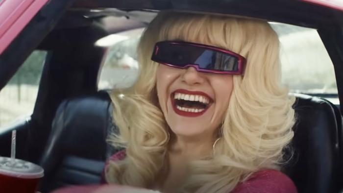 A young woman with platinum blonde hair and sunglasses on cracks a huge smile, pictured from the shoulders up through the driver's side window of a pi