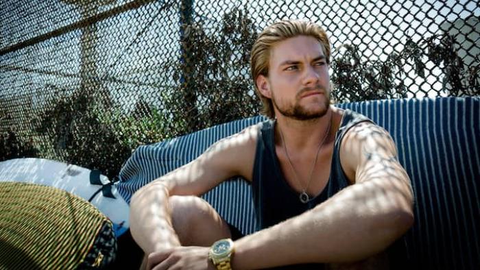 Animal Kingdom TV Show: A blond man wearing a tank sits on the ground in front of a chain-link fence