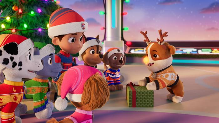 A PAW Patrol Christmas TV show screenshot 1: Ryder and puppies are in front of a Christmas tree watching Rubble hold a green gift box, nose lit.