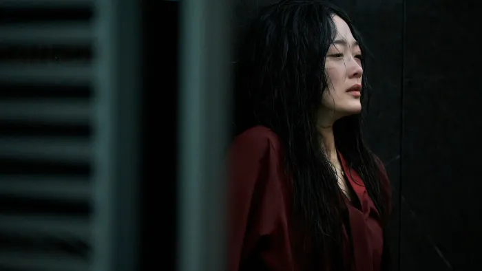 A Korean woman with wet hair and some visible wounds under her red blouse, stands with her shoulders high and her back against the wall