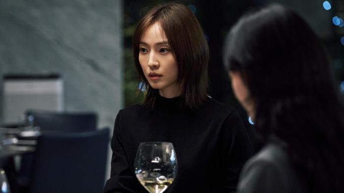 A Korean woman with shoulder-length brown hair sits in a darkened restaurant with a glass of white wine in front of her looking at someone off camera 