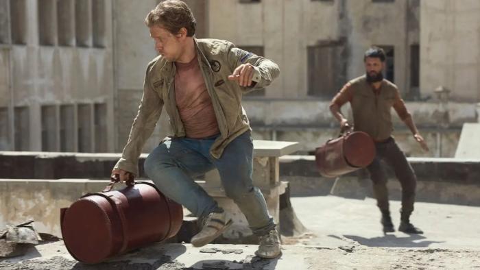 Atomic TV show screenshot 3: Alfie Allen and Shazad Latif run across a building rooftop. They both carry red leather bags shaped like cylinders.