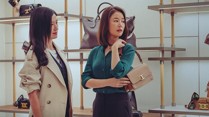 At the Moment TV show screenshot 3: Two women stand in a boutique, the woman on the right models a tan handbag.
