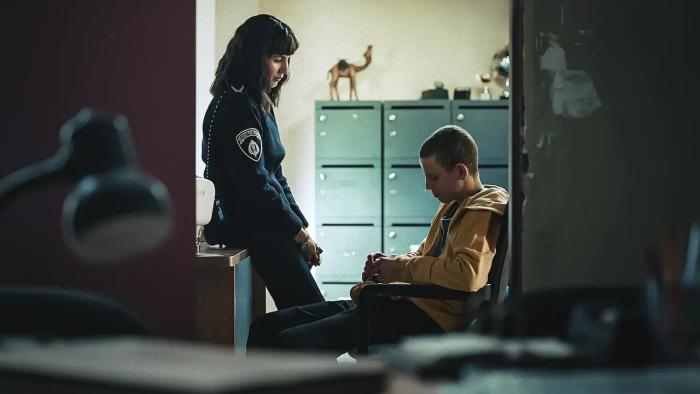 Liraz Chamami leans against a desk in an office. Guy Manster sits on a chair in front of her. He wears a yellow zipped hoodie.