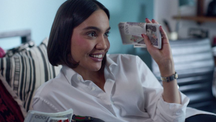 Barracuda Queens screenshot 2: Amina in white shirt lies on bed and holds up wad of cash. 