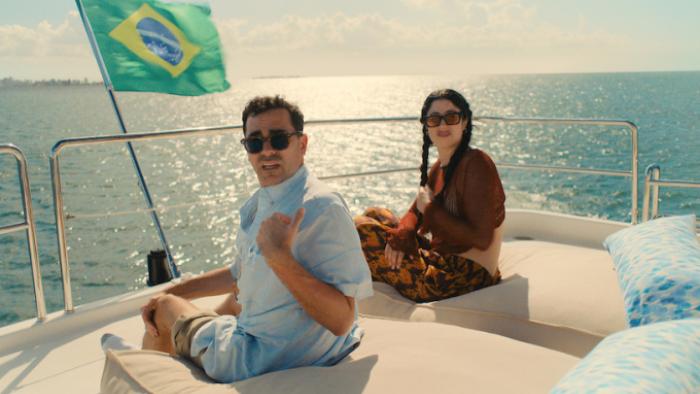 Big Mistakes: Morgan and Nicky sit on a large boat on a waterway; the flag of Brazil flies and Nicky holds a beer; both are looking back with unease