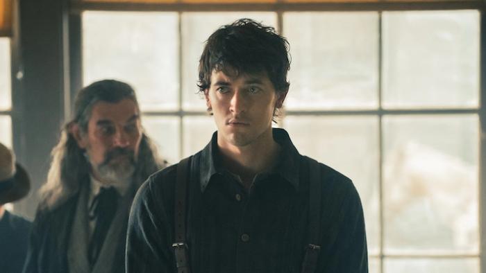 A brown-haired man in a dark shirt and suspenders stands in front of a bright window, he appears to be listening to someone with a concerned look on his face. Two people stand behind him, one a man with a beard and long grey hair and another in a hat, with their face obscured.