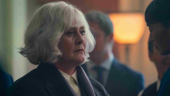 Black Doves TV show screenshot 2: Sarah Lancashire looks amongst a crowd at a party. She wears a white collared shirt, blue coat, and purple scarf.