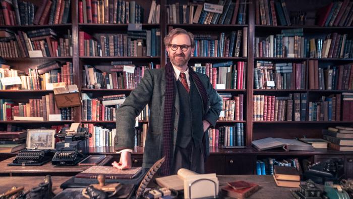 Bookish TV show screenshot 1 :Mark Gatiss stands in front of a cluttered bookshelf. He wears a green suit, red tie, white shirt, and a long scarf.