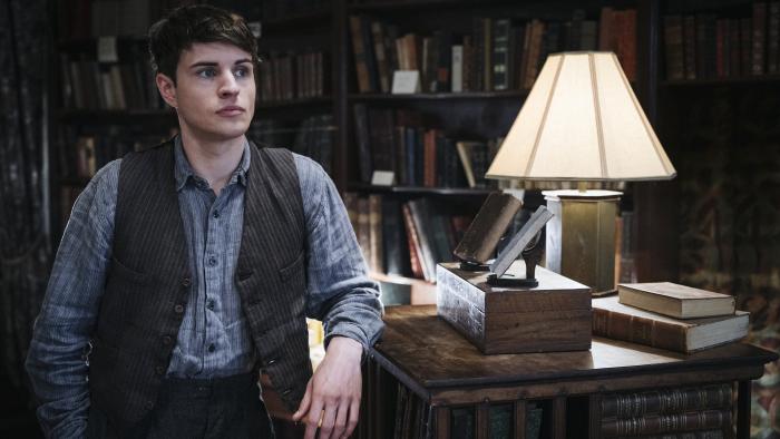 Bookish TV show screenshot 2: Connor Finch leans against a shelf in a bookstore. He wears a blue striped shirt, black striped vest, and dark pants.