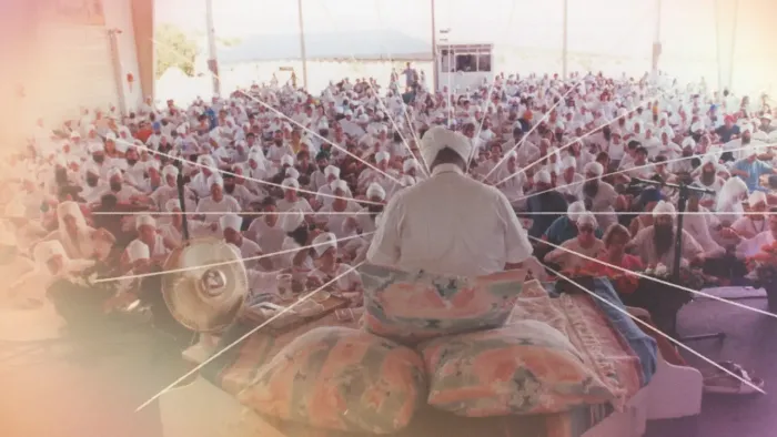 The back of a dark skinned man in white sitting cross legged in a white headwrap in front of a crowd. Drawn lines radiate off of the man to the crowd