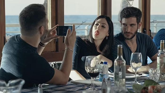 Burning Body TV show screenshot 1: A man snaps a photo of a dark-haired woman sticking her tongue out, another man sits just next to her at a dining table strewn with wine glasses and plates.