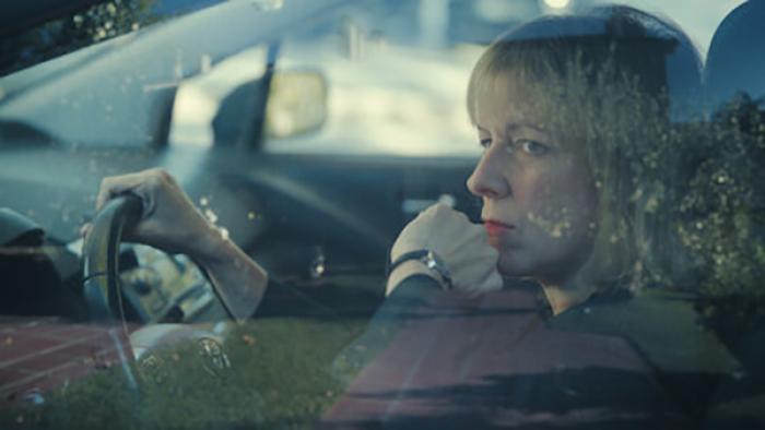 Burning Body TV show screenshot 3: A blonde woman with bangs sits inside a car, hand on the steering wheel, looking out her window with a thoughtful expression on her face.