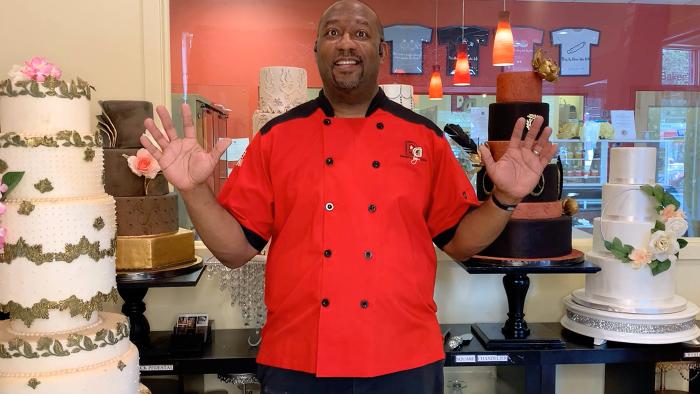 Cake Toppers: A Black man in a red chef's uniform stands smiling with his hands up next to a wedding cake.