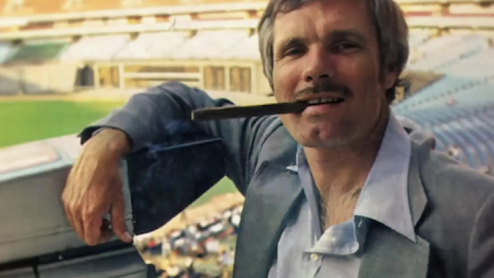 Call Me Ted TV show screenshot 1: A younger Ted Turner smokes a cigar standing on baseball field bleachers.
