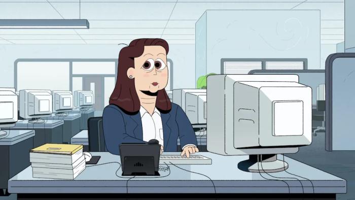 A brunette woman in business casual attire sits at a desk in a bland office setting.