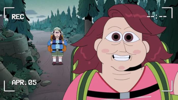 A smiling woman with auburn hair pictured in a video camera's frame. Carol stands on a hiking path in the forest behind her.