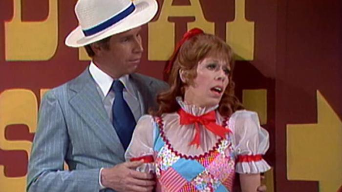 The Carol Burnett Show screenshot 1: Two actors from the Carol Burnett show appear in a skit. One is a man in a grey suit and white hat, the other is a redheaded woman in a colorful red, white, and blue dress.