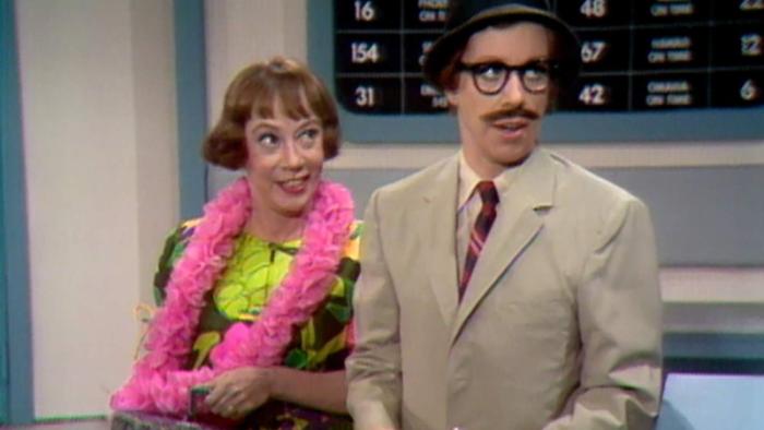 The Carol Burnett Show screenshot 3: Two actors from the Carol Burnett show appear in a skit. A redheaded woman at left holds a bag and is wearing a hot pink scarf. At right, actress Carol Burnett is dressed as a man with a black wig, mustache, and khaki suit.
