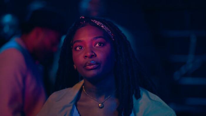 Déja J. Bowens stands in a crowd, watching a performance on stage. She is case in blue and pink light.