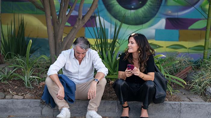 Chivalry TV show screenshot 3: Cameron and Bobby sit on a curb, a colorful mural behind them. Cameron bows his head, Bobby holds her phone and smiles