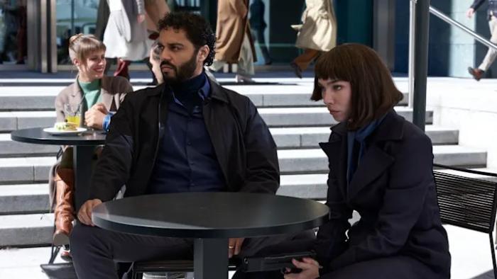 (From R-L) Marouane Zotti and Matilda De Angelis sit at a table. Angelis holds a pistol between her legs, aiming it forward.